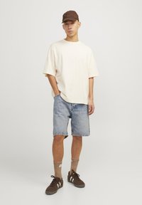 Beige, rövid ujjú póló, világoskék farmer rövidnadrág, barna sportcipők fehér csíkokkal, és barna sapka. A zoknik világosbarna csíkosak.