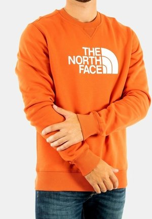 Sudadera naranja de tejido suave, con un diseño de logo blanco y puños acanalados. El diseño es de cuello redondo clásico con un corte relajado.