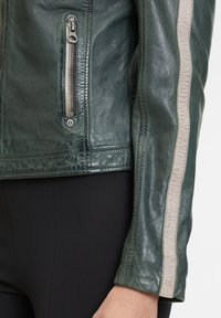 Grüne Lederjacke mit strukturierter Oberfläche, ausgestattet mit einem metallischen Reißverschluss, einer seitlichen Reißverschlusstasche und einem kontrastierenden beigen Streifen entlang des Ärmels.