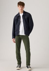 Denim jacket in dark blue with orange stitching, white t-shirt, green pants, and black sneakers with white soles. Model is standing and smiling.