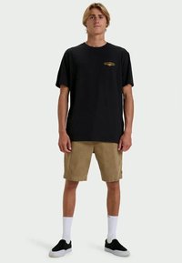 Young man standing, wearing a black t-shirt with small logo, tan shorts, white socks, and black slip-on shoes, against a plain background.