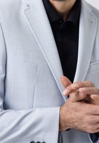 Jasnoszary żakiet o teksturowanej tkaninie, z zapięciem na jeden guzik, z klapami w kształcie wyłogów i kieszenią; noszony na czarnej koszuli z kołnierzykiem.
