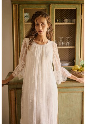 Joven mujer con cabello rizado, vestida con un vestido blanco de encaje, de pie con los brazos apoyados en un armario rústico verde con platos y una cesta de limones.