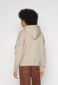 Felpa beige con cappuccio regolabile, tessuto strutturato e dettagli con tasche laterali. Abbinata a pantaloni marroni con cuciture a contrasto.