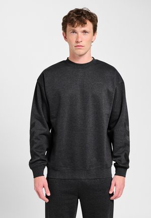 Młody mężczyzna ubrany w ciemnoszarą bluzę z długim rękawem ozdobioną subtelnymi wzorami w kształcie zygzaka na rękawach, stojący na jasnym tle.