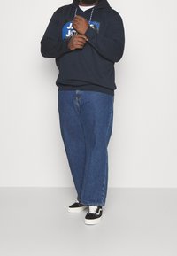 Felpa blu marina con logo sul davanti, abbinata a jeans in denim blu a taglio comodo e sneakers nere con suole e lacci bianchi.