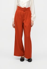 Παντελόνια με φαρδιά μπατζάκια σε ζωηρό κόκκινο ύφασμα. Διαθέτουν ψηλή μέση με ζώνη και πιέτες, συνδυασμένα με μαύρα μοκασίνια για αντίθεση.