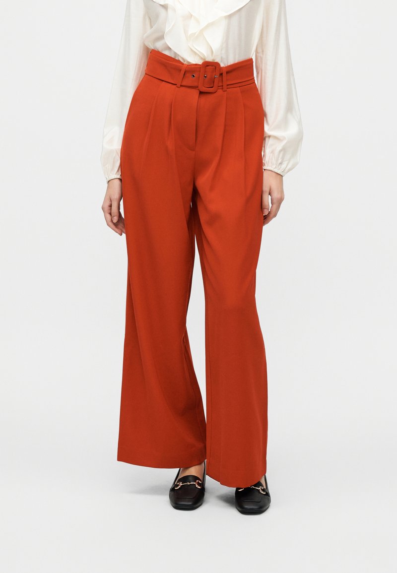 Παντελόνια με φαρδιά μπατζάκια σε ζωηρό κόκκινο ύφασμα. Διαθέτουν ψηλή μέση με ζώνη και πιέτες, συνδυασμένα με μαύρα μοκασίνια για αντίθεση.