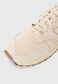 Beige spordijalats, millel on nahast pealis, tekstureeritud paelad ja kummist välispind, millel on minimalistlik disain ja kerge õmblustäide.