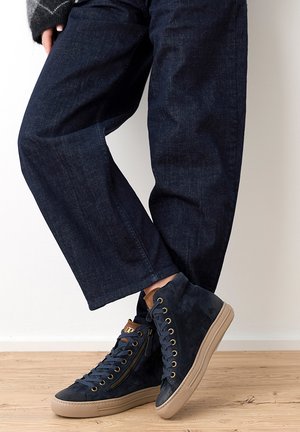 Baskets montantes en daim bleu foncé avec semelles beige, œillets dorés, fermeture éclair latérale ; portées avec un pantalon large en denim foncé sur un sol en bois.