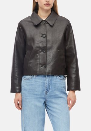 Frau trägt eine dunkelbraune, knöpfbare Lederjacke mit weitem Kragen und hellblaue, hoch taillierte Jeans vor einem einfarbigen Hintergrund.