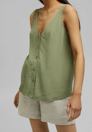 Frau trägt ärmelloses, olivgrünes, geknöpftes Oberteil und beigefarbene Shorts, steht vor einem schlichten grauen Hintergrund.