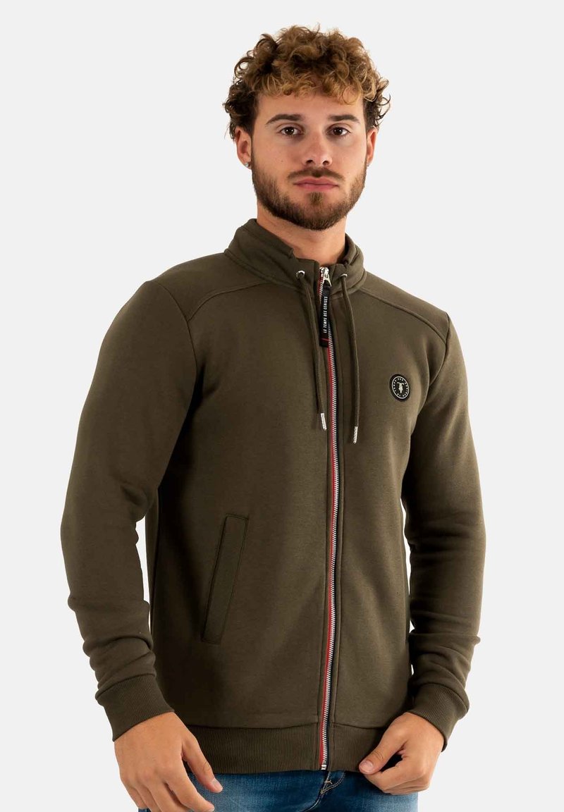 Olijfgroene ritsvest met hoge kraag, voorzakken en een contrasterende rits. Voorzien van ribgebreide boorden en een logo op de borst.