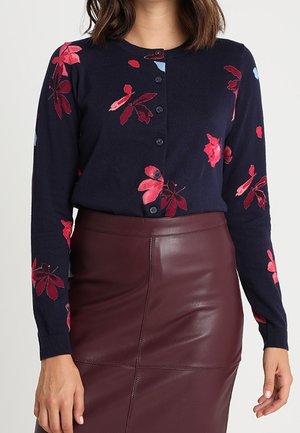 Kobieta ubrana w granatowy rozpinany sweter z czerwonymi kwiatowymi wzorami oraz wysoki, bordowy skórzany spódnicę.