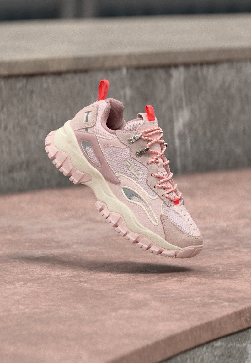 Massiver pink-weißer FILA-Sneaker mit roten Zuglaschen, robuster Sohle und gemusterten Schnürsenkeln, der über einem rötlich-grauen Steinpflaster und Betonstufen schwebt.