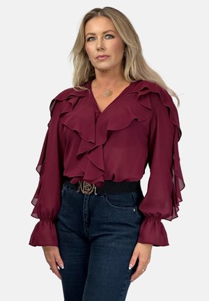 Maroon blouse with ruffled details along the neckline and sleeves, paired with high-waisted dark jeans and a wide belt featuring a logo buckle.