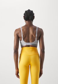 Sportsbh i grått med justerbare stropper som har logo-detaler, sammen med høyhårede gule leggings med en glatt tekstur.