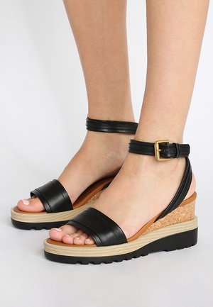 Pies que llevan sandalias cuña de cuero negro con tira en el tobillo, tacones de corcho y suelas beige sobre un fondo claro y liso.