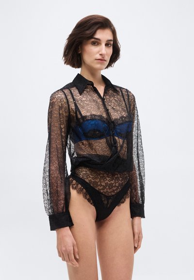 Kvinde med kort mørkt hår iført en sort blonder langærmet skjorte over en blå bh og matchende sort blonderundertøj mod en ensfarvet baggrund.