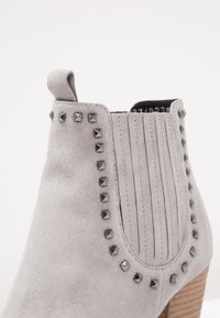 Bottine en daim gris clair avec des clous carrés noirs délimitant le panneau élastique latéral et un talon en bois empilé.