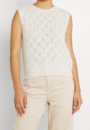 Mouwloos wit gebreid topje met textuur van een diamantpatroon, gedragen met lichtbeige broek, getoond van hals tot heupen.