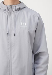 Lichtgrijze ritsjas met capuchon, gemaakt van een glad, lichtgewicht materiaal. Heeft een wit logo op de borst en verstelbare trekkoorden.