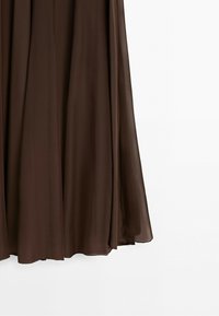 Vloeiend bruin chiffon drapeert in zachte plooien tegen een eenvoudige witte achtergrond en toont een deel van een lange rok of jurk.
