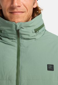 Giacca verde chiaro con colletto alto, chiusura con zip e patch logo nero. Il materiale sembra liscio con una leggera lucentezza. La zip presenta dettagli a contrasto.