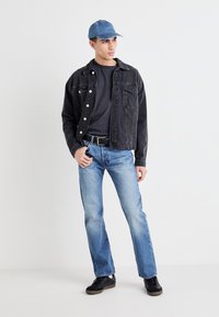 Chaqueta de mezclilla negra, camiseta gris, jeans azules y zapatos negros. Una gorra azul claro completa el atuendo casual con un ajuste relajado.