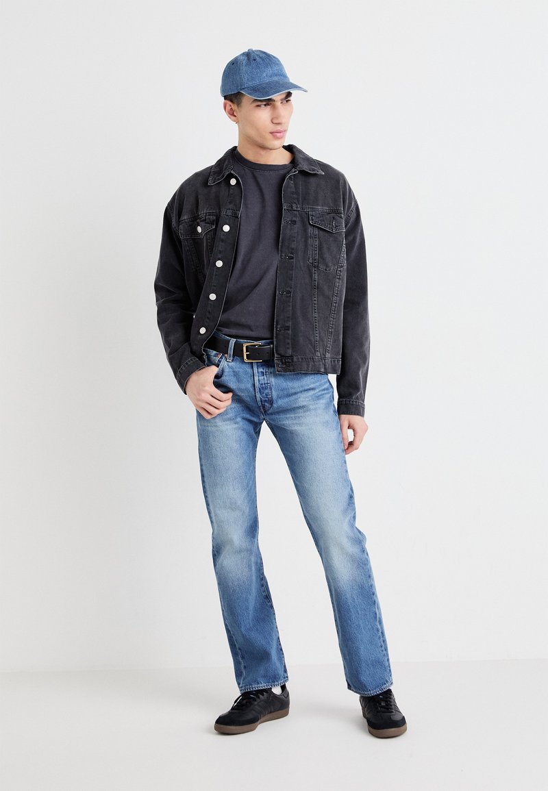 Sort denimjakke, grå t-shirt, blå jeans og sorte sko. En lys blå kasket fuldender det afslappede outfit med en løs pasform.