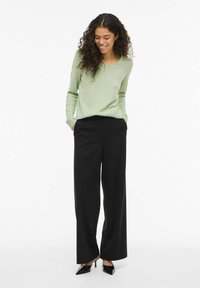 Femme aux cheveux bouclés portant un pull vert clair, un pantalon noir à jambes larges et des escarpins noirs à bouts pointus, debout les mains dans les poches.