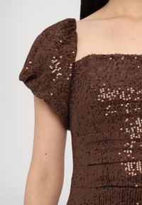 Gros plan d'une personne portant une robe à sequins marron à manches courtes avec des épaules bouffantes et un décolleté carré.