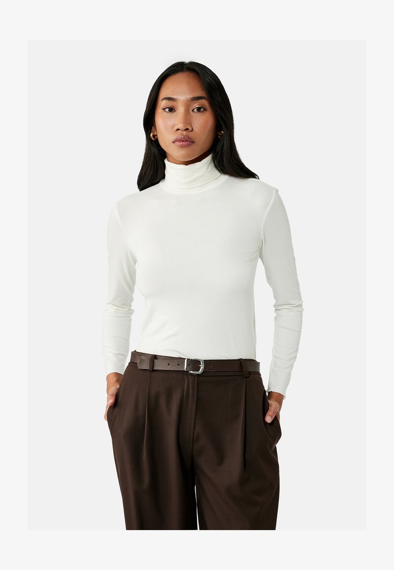 Kobieta o długich ciemnych włosach, ubrana w biały golf i brązowe spodnie z wysokim stanem i paskiem, stojąca z rękami w kieszeniach na białym tle.