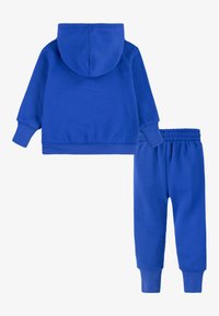 Zils džemperis ar kapuci, kenguru kabatu un elastīgiem aprocēm, savienots ar atbilstošām zilām sporta biksēm ar elastīgu jostu un potīšu aprocēm.