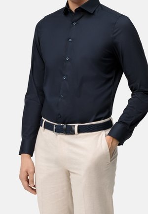 Homme portant une chemise bleu marine foncé à manches longues boutonnée, un pantalon beige et une ceinture bleu marine tissée, debout avec une main dans la poche.