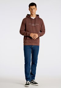 Brun huvtröja med vitt logotyp, med dragsko i huvan och ribbade ärmslut, bärs över en vit skjorta. Kombineras med blå jeans och svarta sneakers.