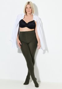 Donna con un bob biondo che indossa un reggiseno nero, pantaloni a vita alta oliva e una camicia bianca aperta, in piedi con le gambe incrociate, su uno sfondo bianco.