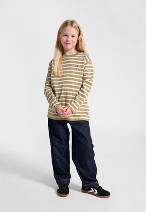 Blond meisje staand, draagt een beige en wit gestreept shirt met lange mouwen, donkerblauwe spijkerbroek en zwarte sneakers met witte chevron strepen.