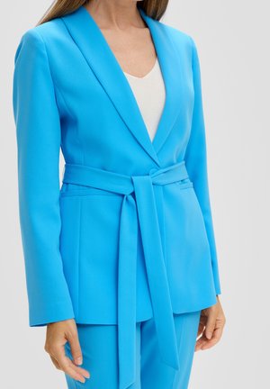 Kvinde iført en klar blå blazer med bælte og matchende bukser over en hvid top, vist fra brystet til hoften.