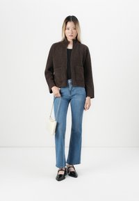 Femme debout portant un cardigan zippé marron, un haut noir, un jean bleu, des talons noirs, et tenant un petit sac à main blanc avec une bandoulière en chaîne.