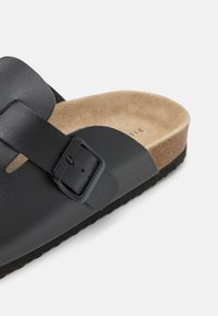 Sandale en cuir noir avec une large sangle et une boucle réglable. La semelle intérieure en liège offre une surface texturée bronzée, complétée par une semelle extérieure en caoutchouc.
