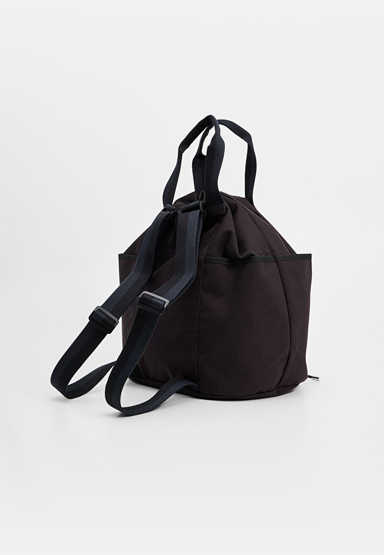 Mochila de tela negra con forma redondeada, que cuenta con dos asas en la parte superior y correas ajustables, acentuada por un cierre con cremallera y bolsillos laterales.