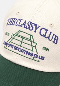 Cappellino da baseball con una corona color crema e visiera verde. Dettagli grafici e testo ricamati in blu e verde. Accenti cuciti visibili.