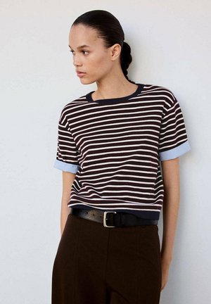 Femme aux cheveux tressés portant un t-shirt à manches courtes rayé bleu marine et blanc, rentré dans un pantalon foncé avec une ceinture noire.