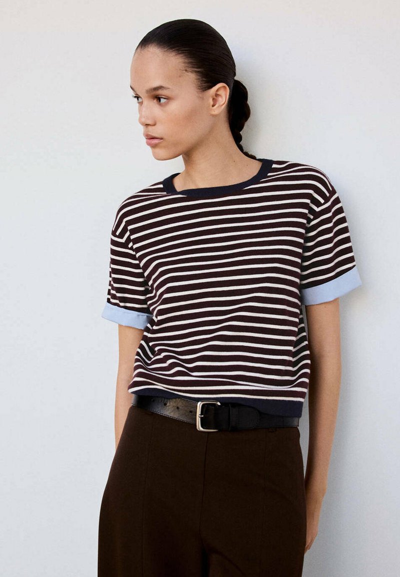 Femme aux cheveux tressés portant un t-shirt à manches courtes rayé bleu marine et blanc, rentré dans un pantalon foncé avec une ceinture noire.