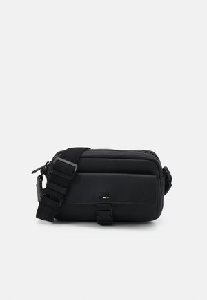 Bolso bandolera de cuero negro con correa ajustable, hebilla frontal y pequeño logo en relieve sobre un fondo claro y liso.