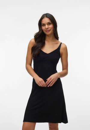 Kvinde med langt mørkt hår iført en ærmeløs sort kjole med justerbare stropper, stående foran en ensfarvet hvid baggrund.