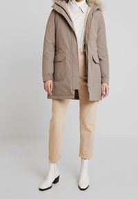 Beige parka-takki turkiksella reunustetulla hupulla, kahdella etutaskulla ja joustavilla hihoilla, yhdistettynä beigeihin housuihin ja valkoisiin nilkkureihin.