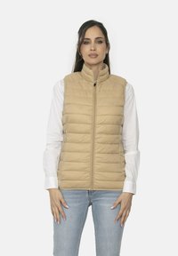 Chaleco acolchado beige con cuello alzado, costuras horizontales y cremallera frontal. Lleva puesto sobre una blusa blanca y combinado con jeans de color azul claro.