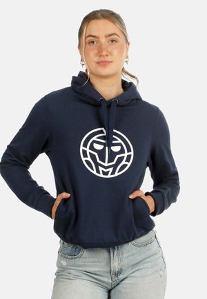 Frau mit hellem Haar, die einen dunkelblauen Hoodie mit weißem geometrischem Gesicht-Logo, Jeans und die Hände in den Taschen des Hoodies trägt.